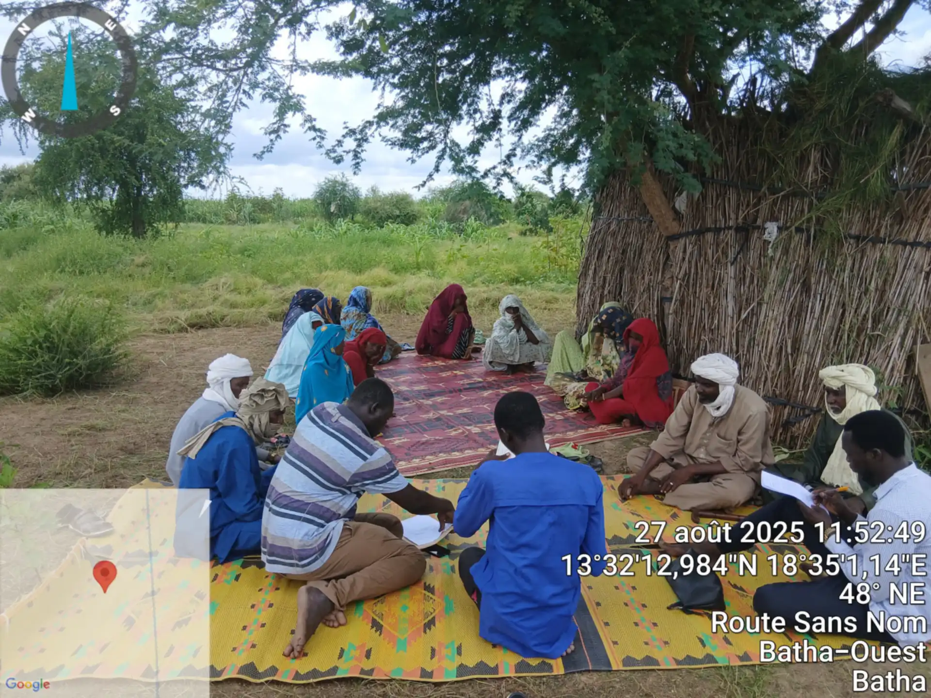 Groupe de discussion mixte assis en cercle sur des nattes, à l’ombre d’un acacia, devant un abri de paille.